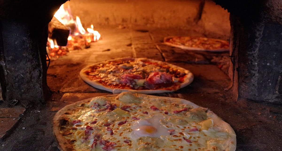 Trois pizzas dans le four à bois, les flammes au fond de la voûte.