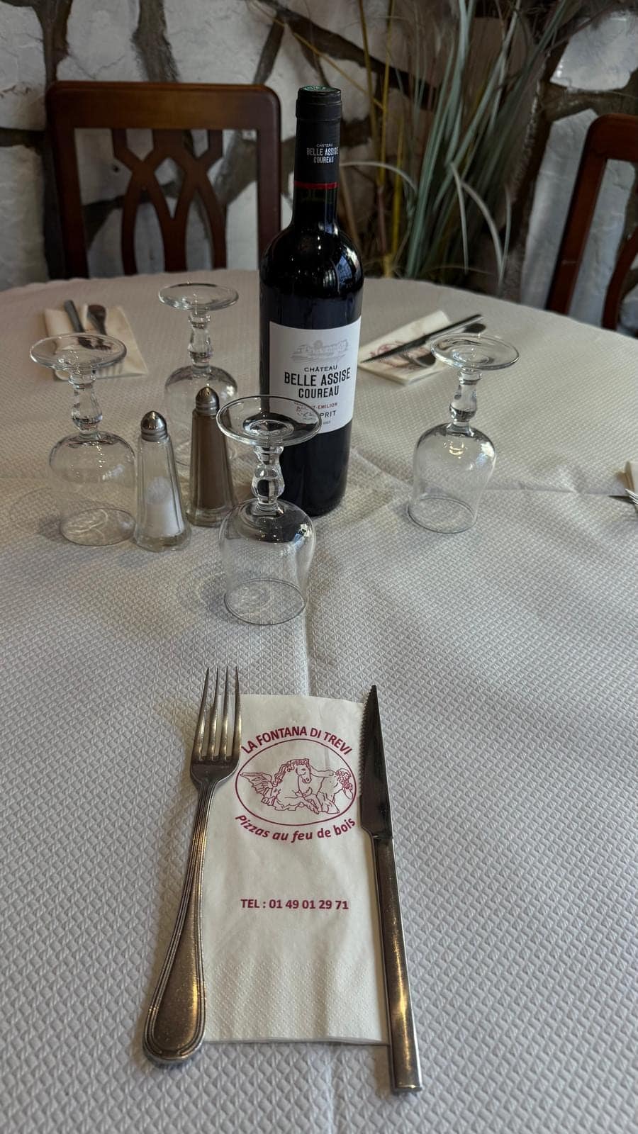 Table ronde dressée : bouteille de vin rouge, verres, couverts et serviette au logo de la maison.