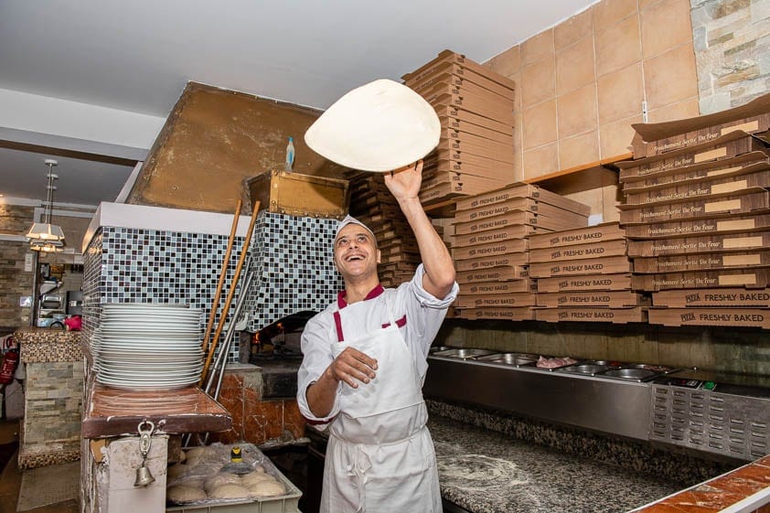 Le pizzaiolo fait tourner la pâte en l’air devant le plan de travail, entre les cartons à pizza.