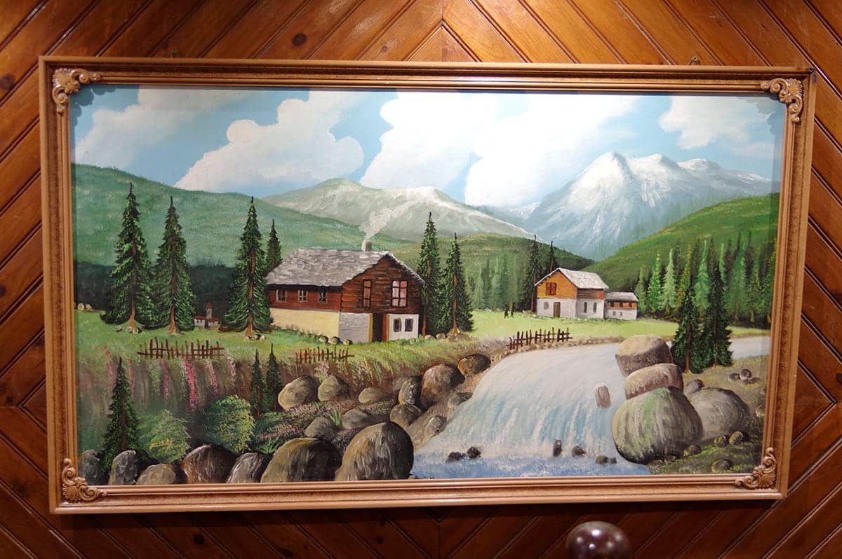 Tableau encadré d’un chalet de montagne au bord d’un torrent, sur un mur lambrissé.